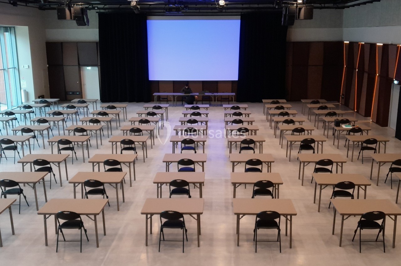 Salle de conférence avec des rangées de tables et chaises alignées face à un écran de projection allumé.