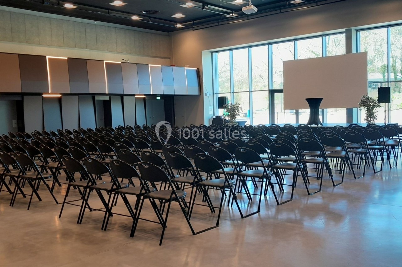 Salle de conférence lumineuse avec des rangées de chaises noires alignées face à un écran blanc.