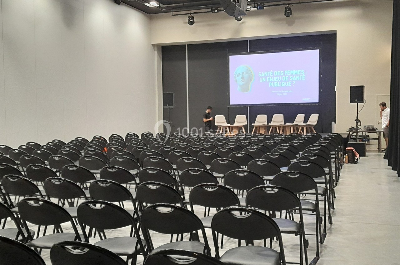 Salle de conférence vide avec rangées de chaises noires alignées face à une scène équipée d'un écran et de chaises blanches.