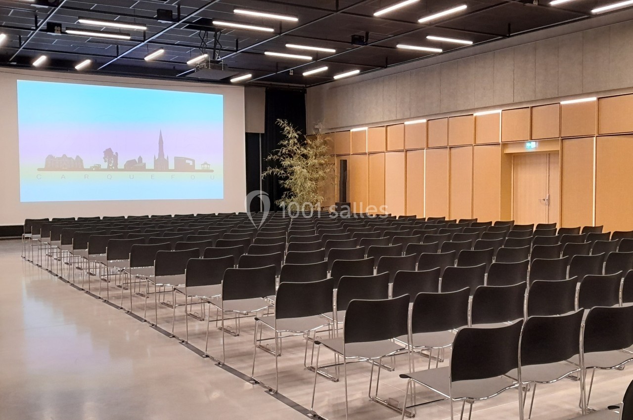 Salle de conférence vide avec des rangées de chaises noires alignées face à un écran de projection allumé.