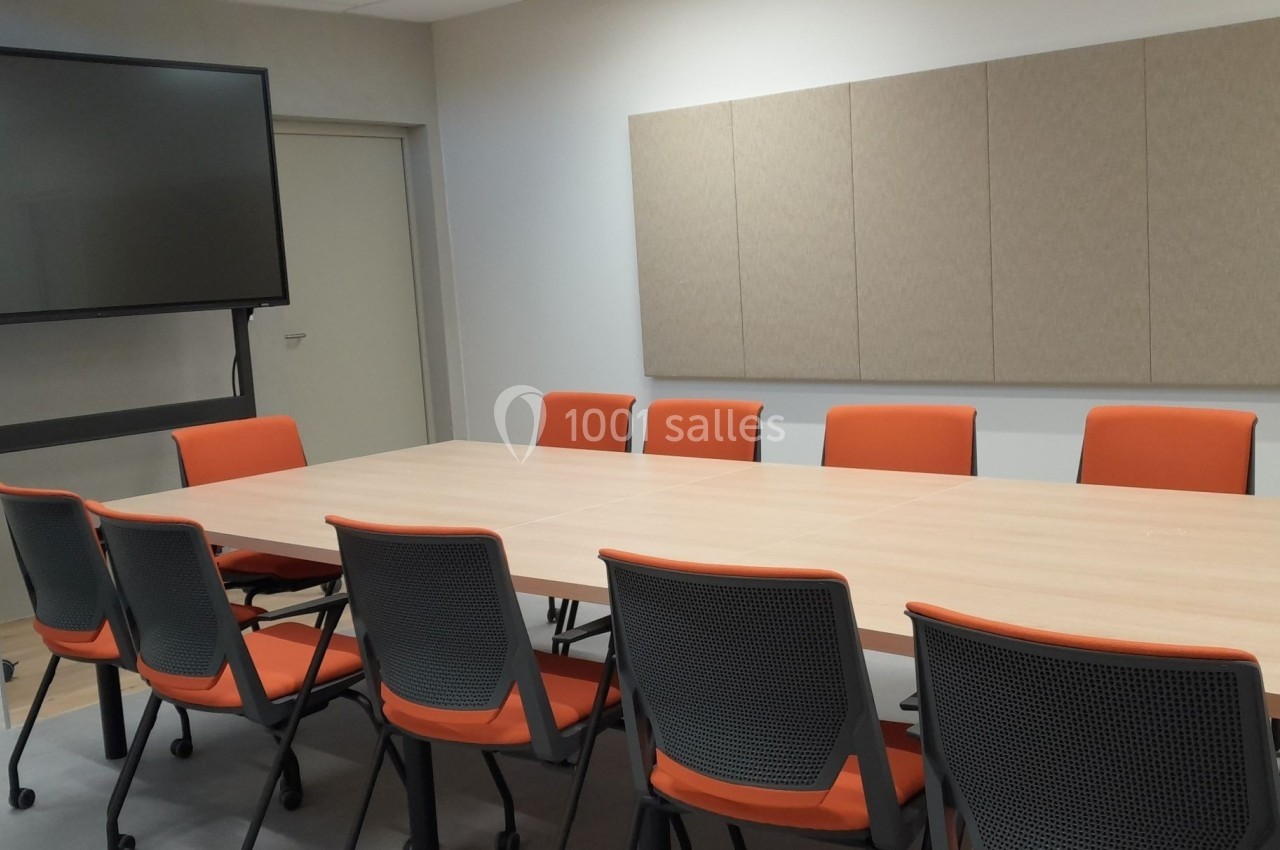 Salle de réunion avec une grande table en bois, des chaises orange et un écran noir monté sur un support.