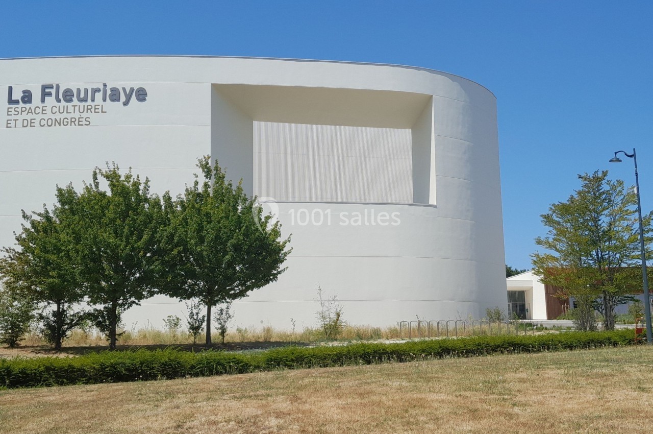 Façade blanche du bâtiment La Fleuraye, espace culturel entouré d'arbres et d'une pelouse sous un ciel bleu.