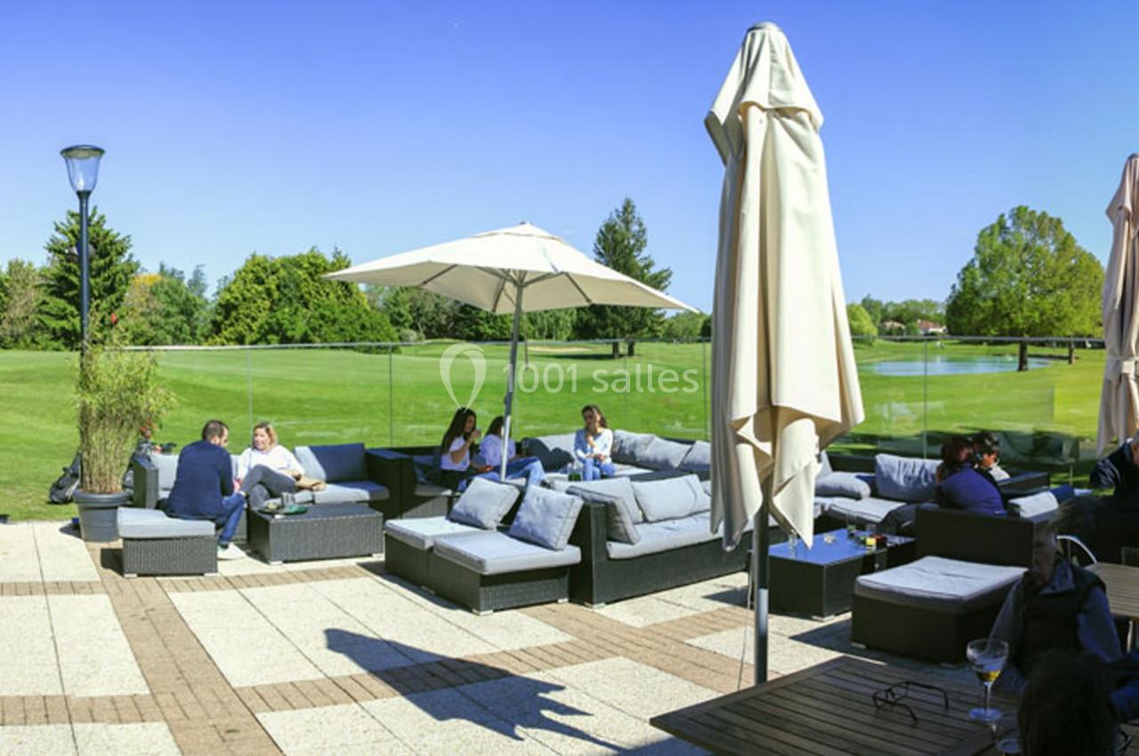 Terrasse avec canapés, parasols et personnes discutant, donnant sur un grand espace vert par une journée ensoleillée.