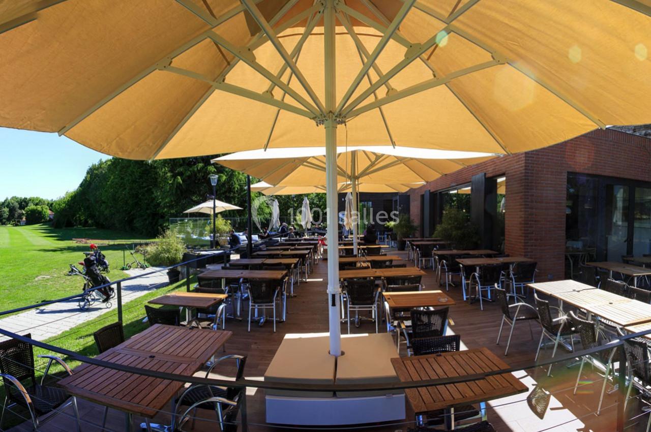 Terrasse extérieure avec tables et chaises sous de grands parasols beige, entourée de verdure et d'un bâtiment en briques.