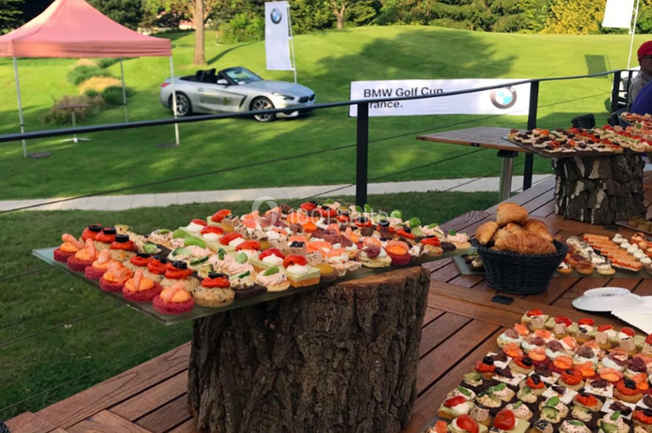 Buffet de canapés variés disposés sur des tables en bois, avec une voiture et un panneau ’BMW Golf Cup France’ en arrière…