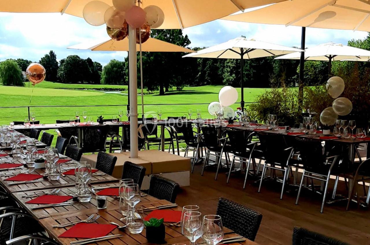 Tables dressées avec nappes rouges et verres, disposées en terrasse ombragée, surplombant un terrain verdoyant.