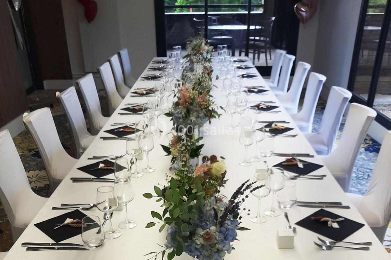 Table élégamment dressée avec nappes blanches, bouquets floraux, verres et serviettes noires dans une salle lumineuse.