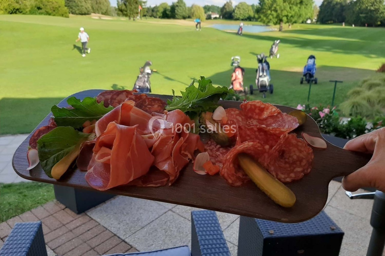 Planche de charcuterie avec cornichons et verdure tenue en avant-plan, vue sur un terrain de golf en arrière-plan.