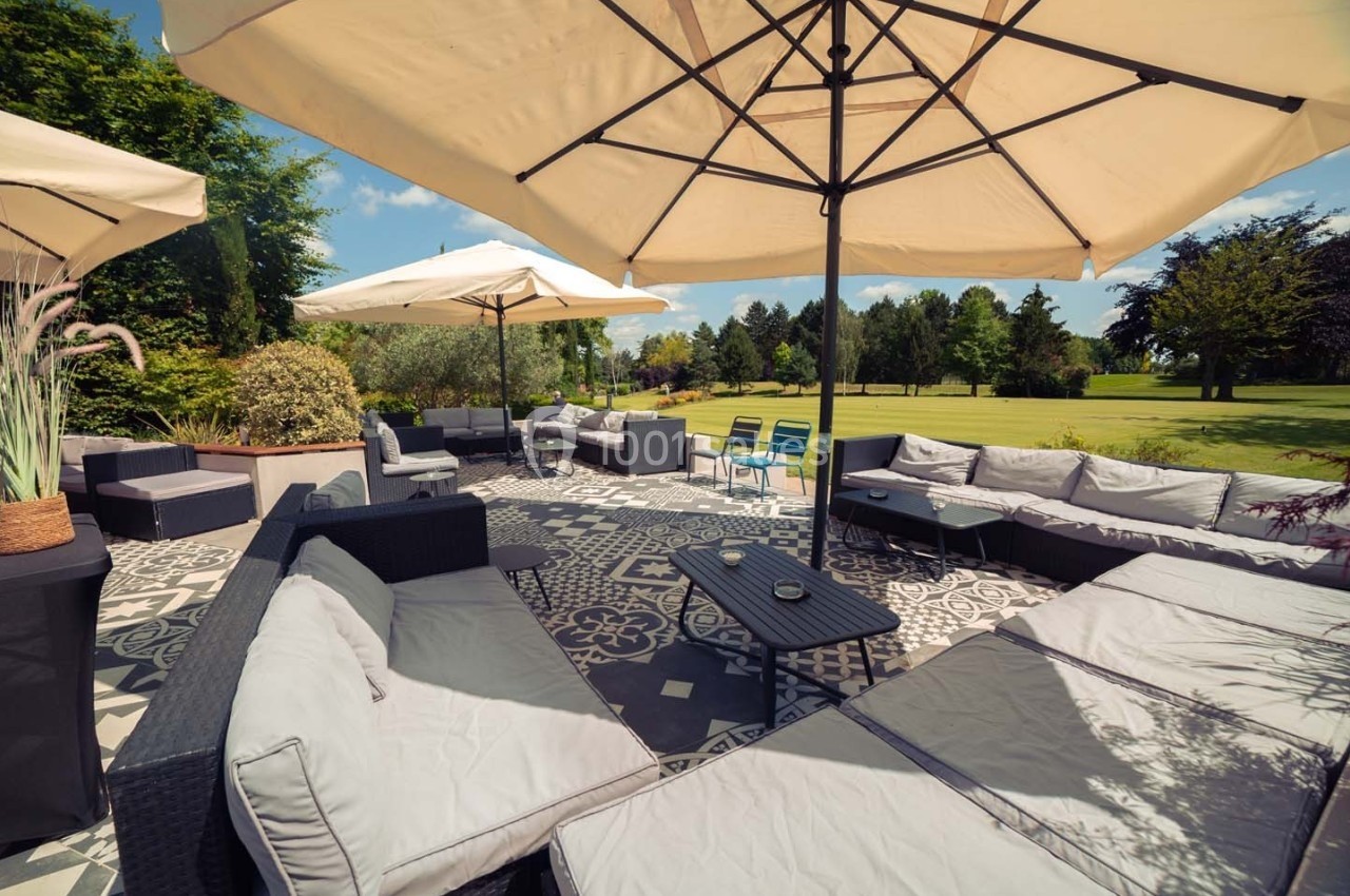 Terrasse aménagée avec canapés, tables basses et parasols, donnant sur un grand jardin verdoyant par temps ensoleillé.