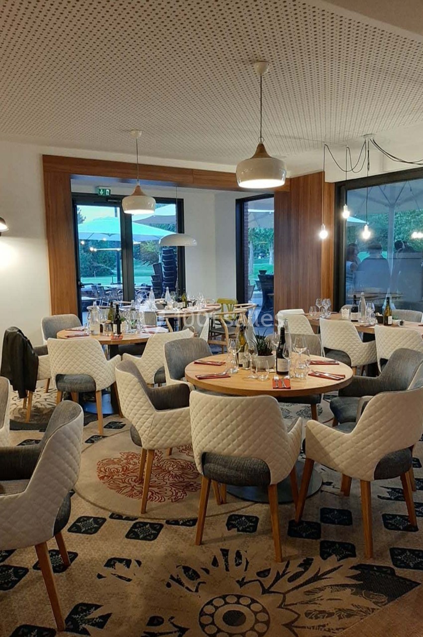 Salle de restaurant moderne avec tables dressées, chaises rembourrées et grandes baies vitrées donnant sur une terrasse.