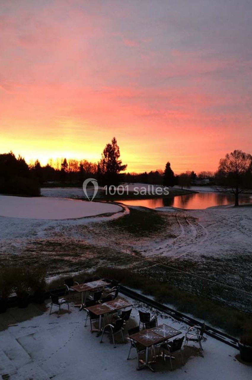 Paysage hivernal avec un terrain enneigé, des tables extérieures et un coucher de soleil vibrant au-dessus d'un lac.