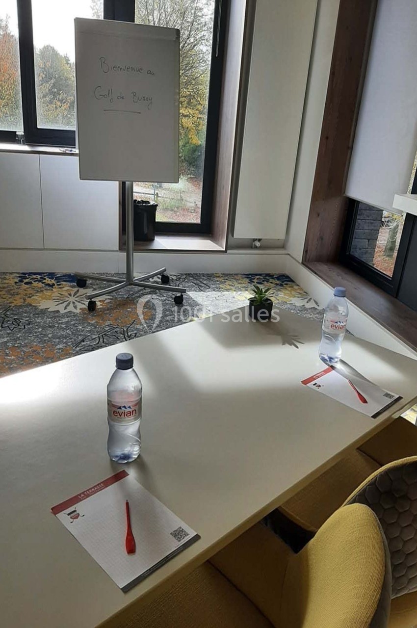 Salle de réunion lumineuse avec table, chaises, bouteilles d'eau, blocs-notes, stylos et tableau blanc près d'une fenêtre.