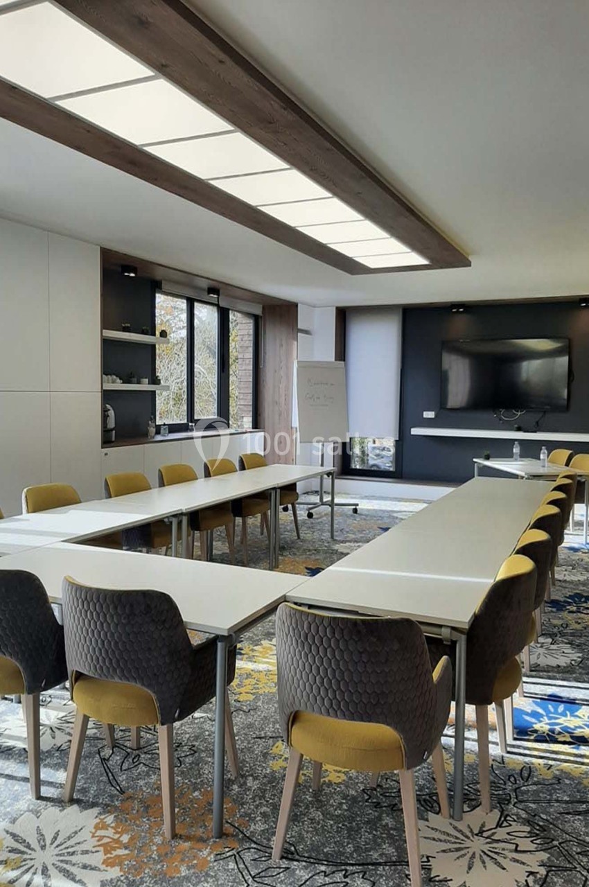 Salle de réunion lumineuse avec tables disposées en U, chaises jaunes et grises, écran mural et tableau blanc.