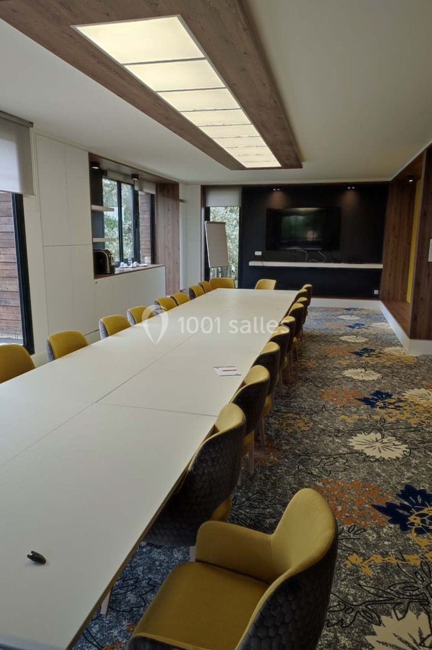 Salle de réunion lumineuse avec une grande table blanche entourée de chaises jaunes et un écran mural au fond.