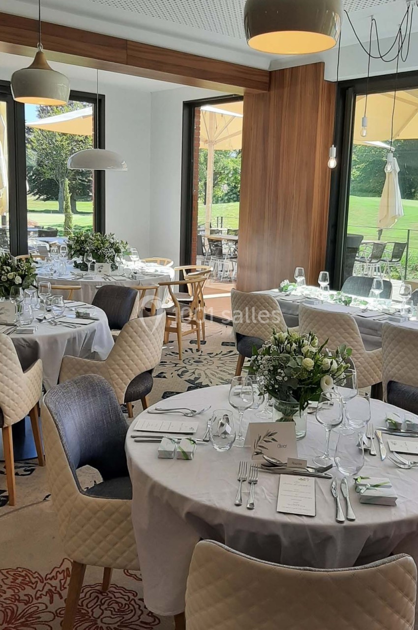 Salle de réception lumineuse avec tables rondes dressées, décorées de fleurs blanches, donnant sur une terrasse et un jardin.