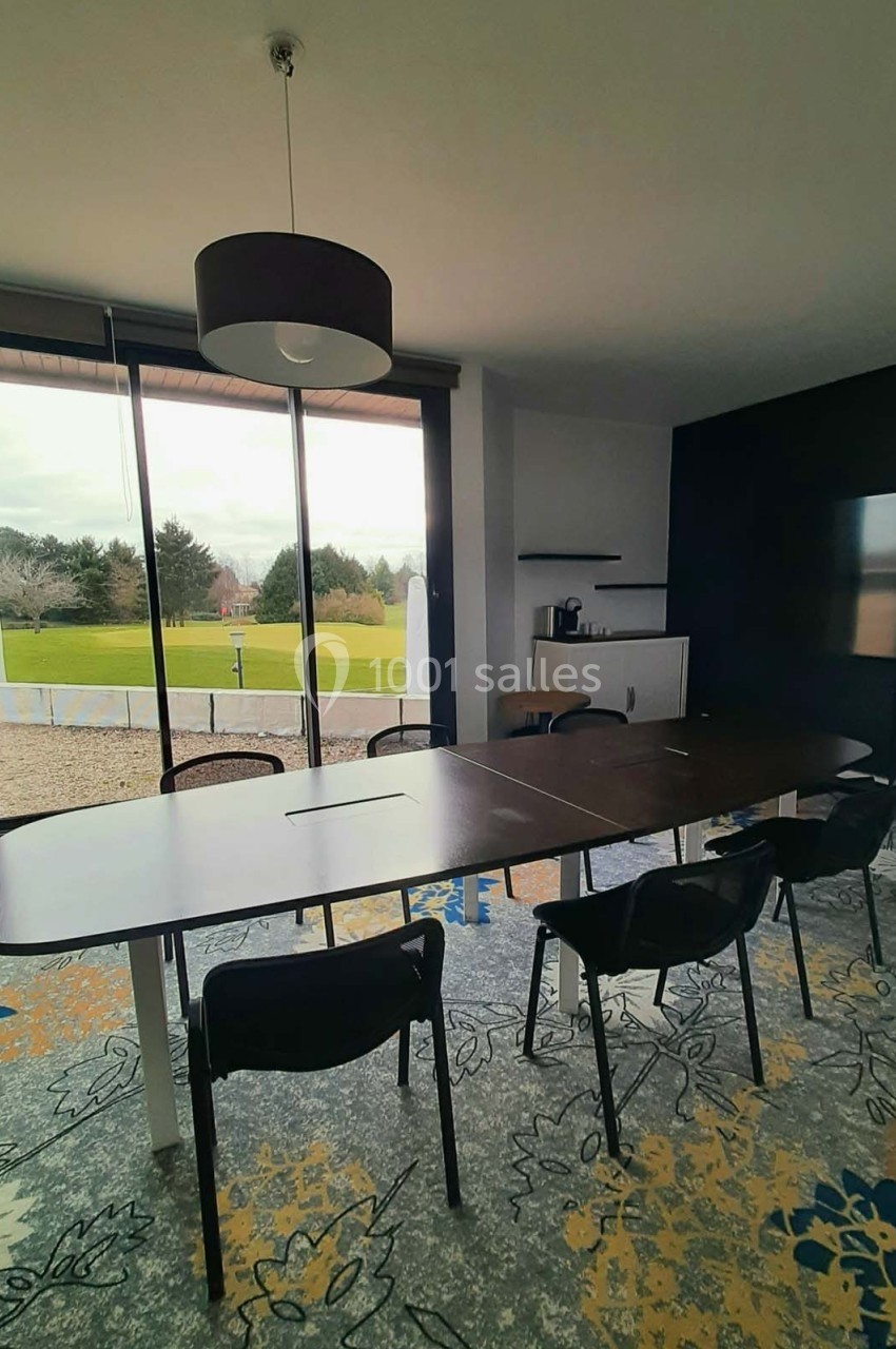 Salle de réunion lumineuse avec table ovale, chaises noires, écran, et vue sur un espace extérieur verdoyant.