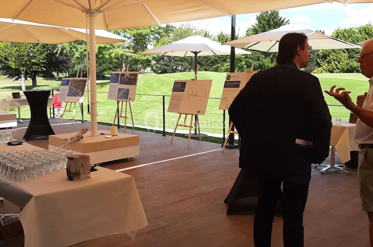 Des personnes discutent sous une terrasse couverte, entourées de panneaux d'exposition et de tables dressées.