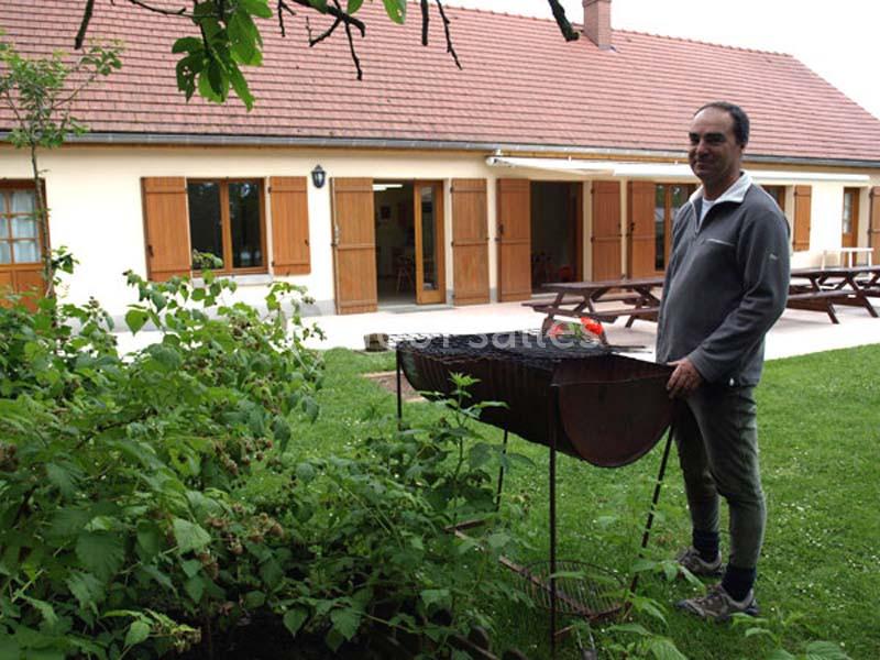 Un homme debout près d'un barbecue dans un jardin, devant une maison avec des volets en bois et des tables de pique-nique.