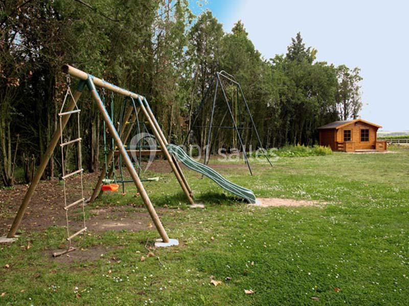 Aire de jeux extérieure avec balançoire, toboggan et cabane en bois, entourée d'herbe et d'arbres.
