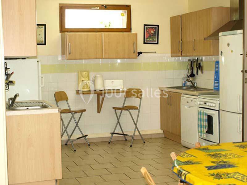 Cuisine équipée avec meubles en bois clair, électroménagers, table pliante avec deux chaises et fenêtre en hauteur.