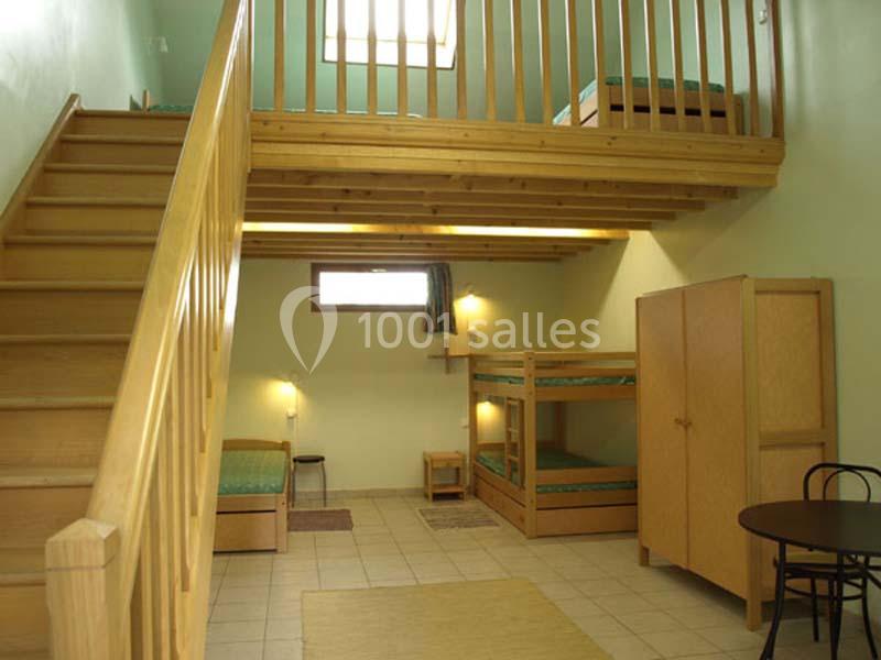 Chambre avec mezzanine en bois, lits superposés, lit simple, armoire, table et chaises, éclairée par une fenêtre.