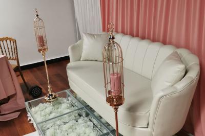 Canapé blanc devant un rideau rose, entouré de bougeoirs dorés en forme de cages et d'une table en verre.