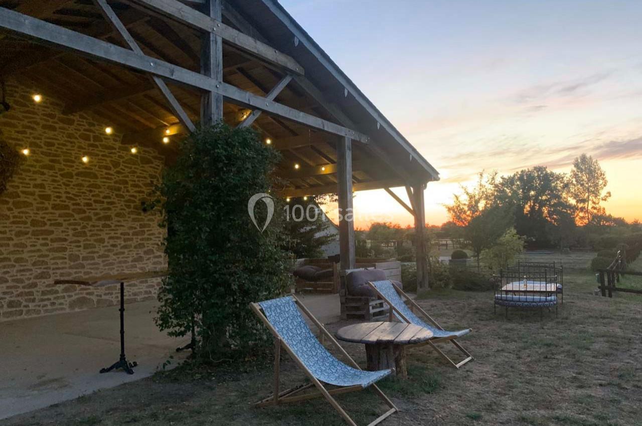Terrasse en bois éclairée au crépuscule, avec chaises longues, table basse et vue sur un jardin arboré.