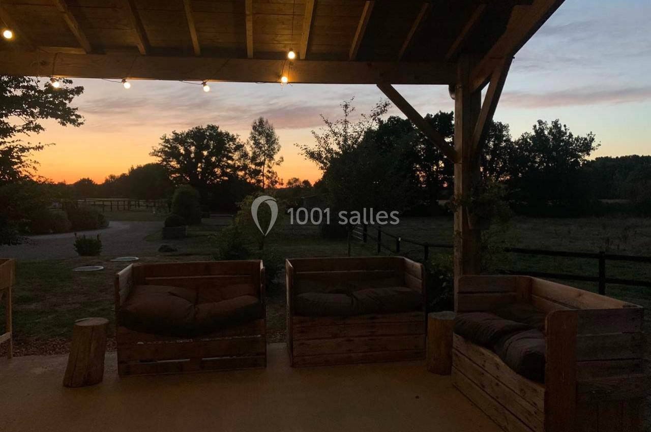 Terrasse en bois avec fauteuils rustiques sous un auvent, éclairée par des guirlandes lumineuses au coucher du soleil.