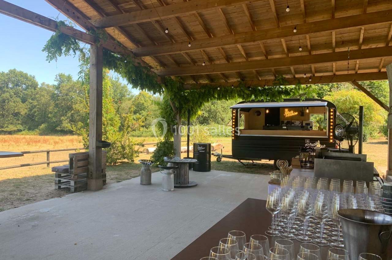 Abri en bois ouvert sur un paysage champêtre, avec un food truck noir et des verres disposés sur une table.