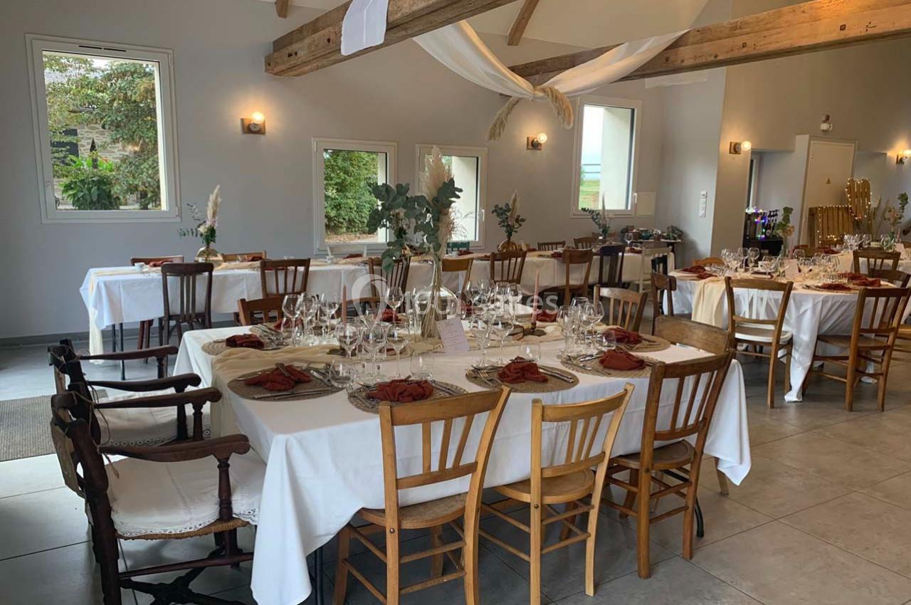 Salle de réception lumineuse avec tables dressées, nappes blanches, chaises dépareillées et décoration florale.