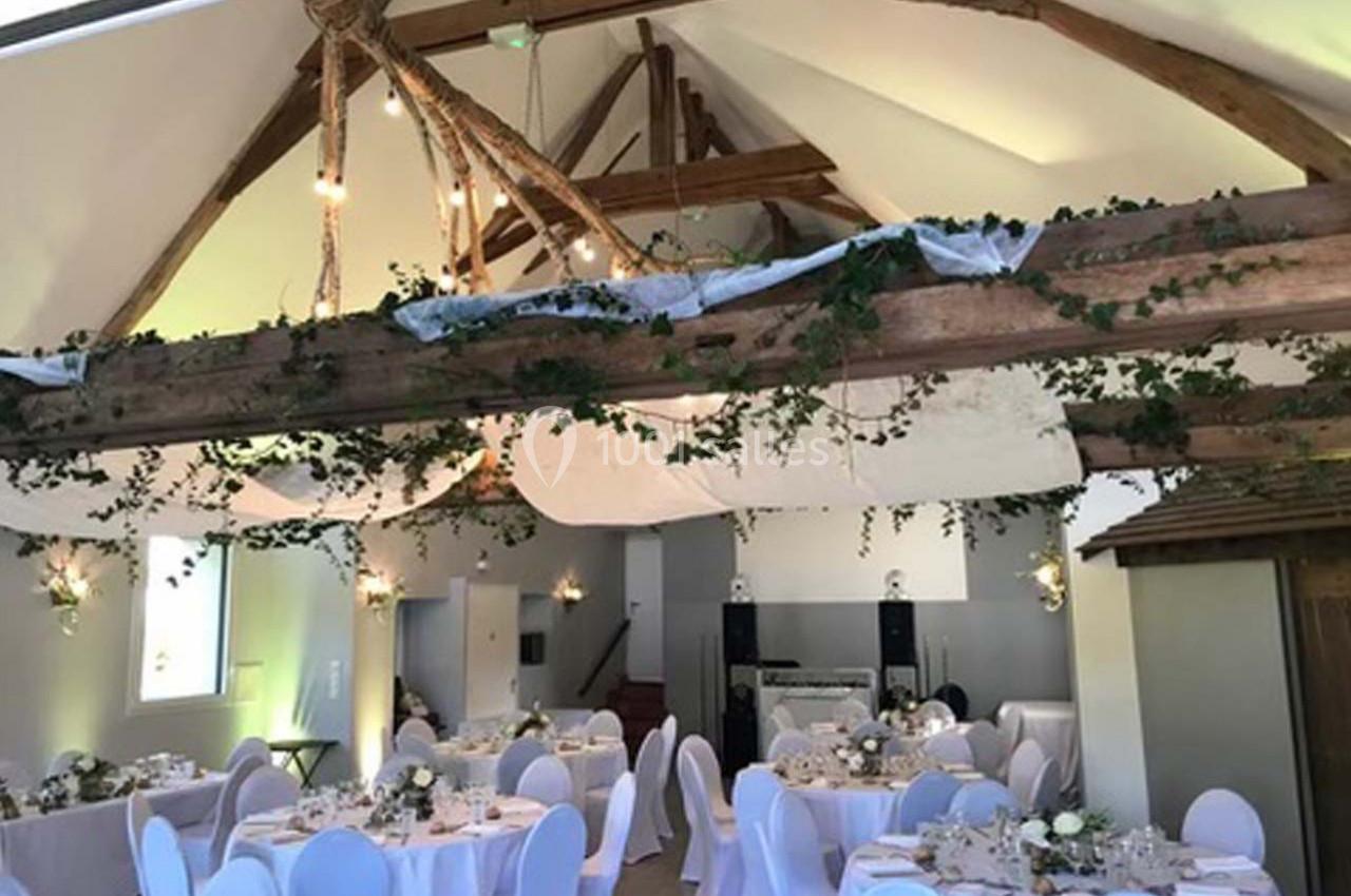 Salle de réception décorée avec des tables rondes dressées, des chaises blanches et des poutres ornées de feuillage.