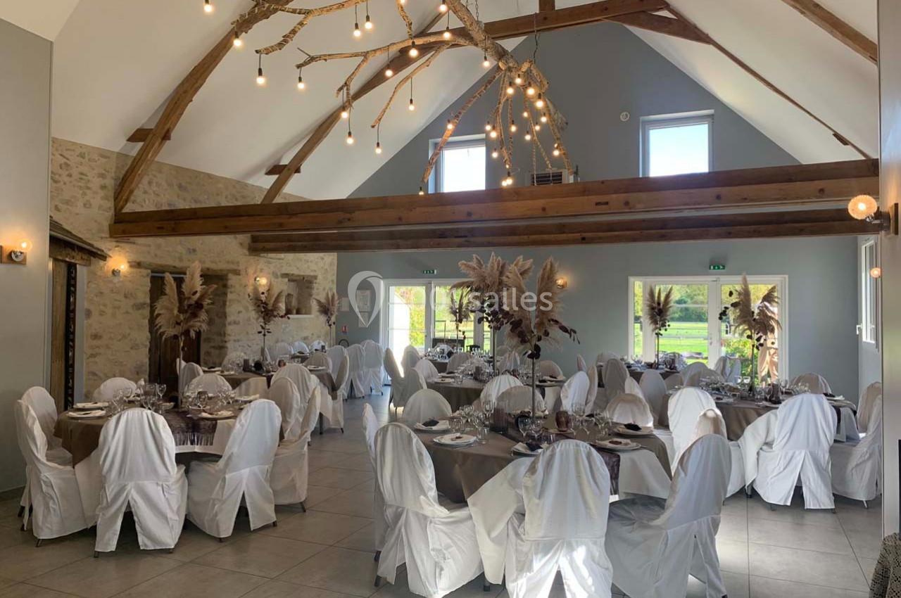 Salle de réception lumineuse avec tables rondes décorées, chaises blanches et poutres apparentes au plafond.