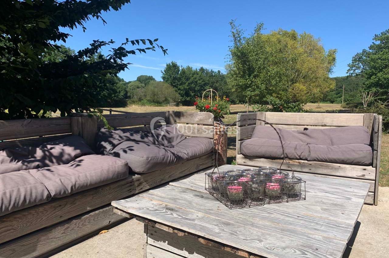 Salon de jardin en bois avec coussins gris, table basse et bougies, situé en extérieur sous un ciel dégagé.