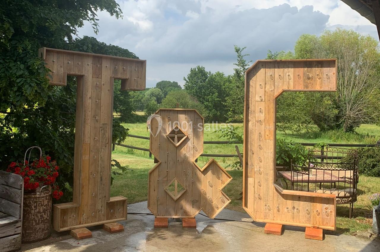 Lettres géantes en bois ’T & C’ disposées en extérieur, devant un paysage verdoyant et un ciel partiellement nuageux.