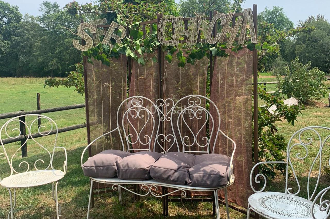 Banc et chaises en fer forgé avec coussins devant un paravent décoré de lettres, dans un jardin verdoyant.