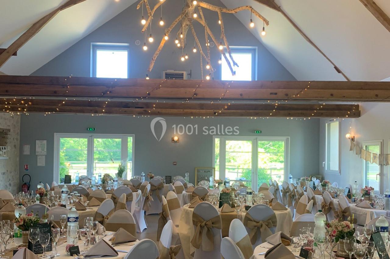 Salle de réception décorée pour un mariage, avec tables dressées, chaises ornées de nœuds et guirlandes lumineuses…