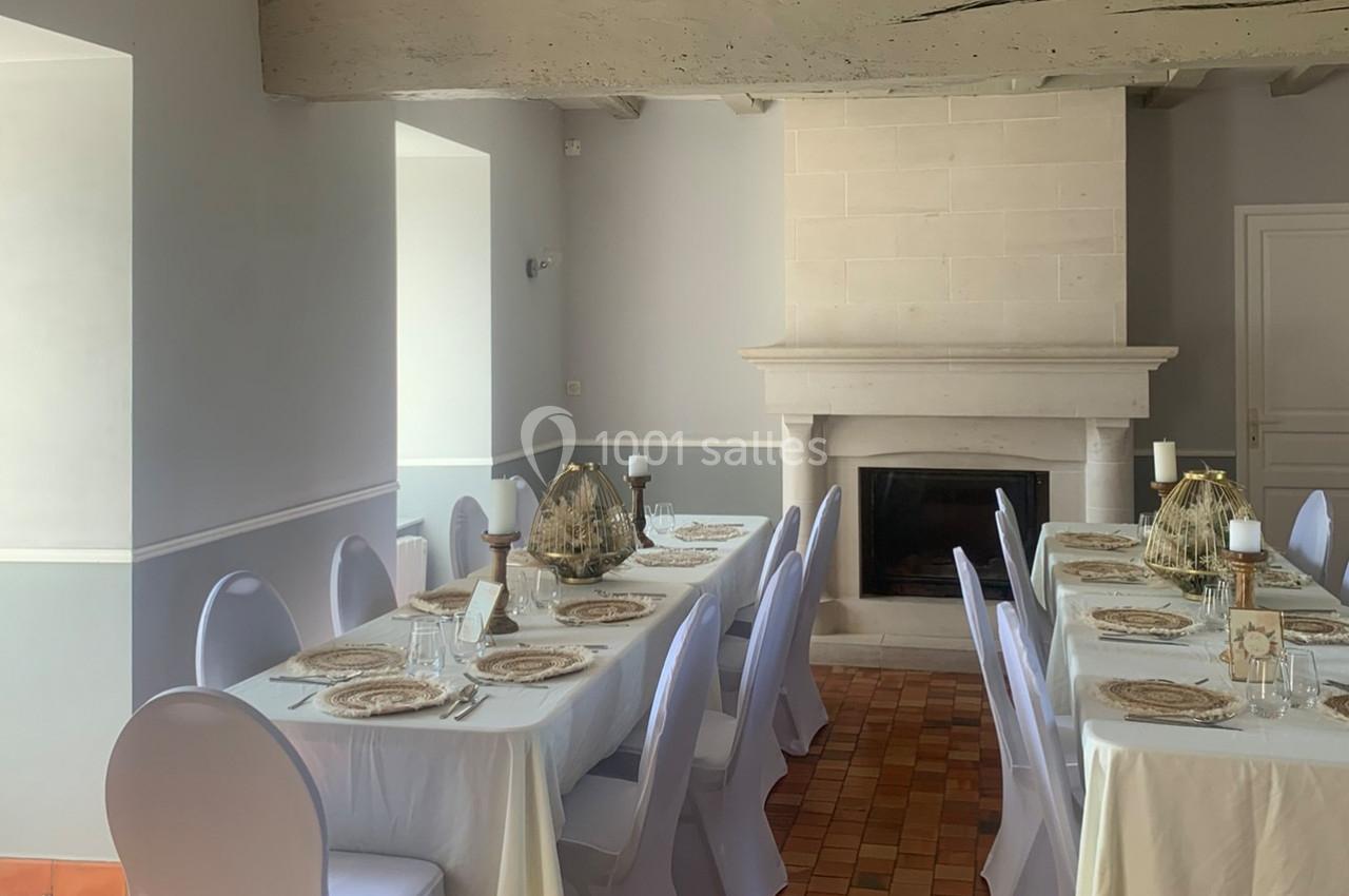 Salle à manger lumineuse avec deux grandes tables dressées, nappes blanches et cheminée en pierre au fond.