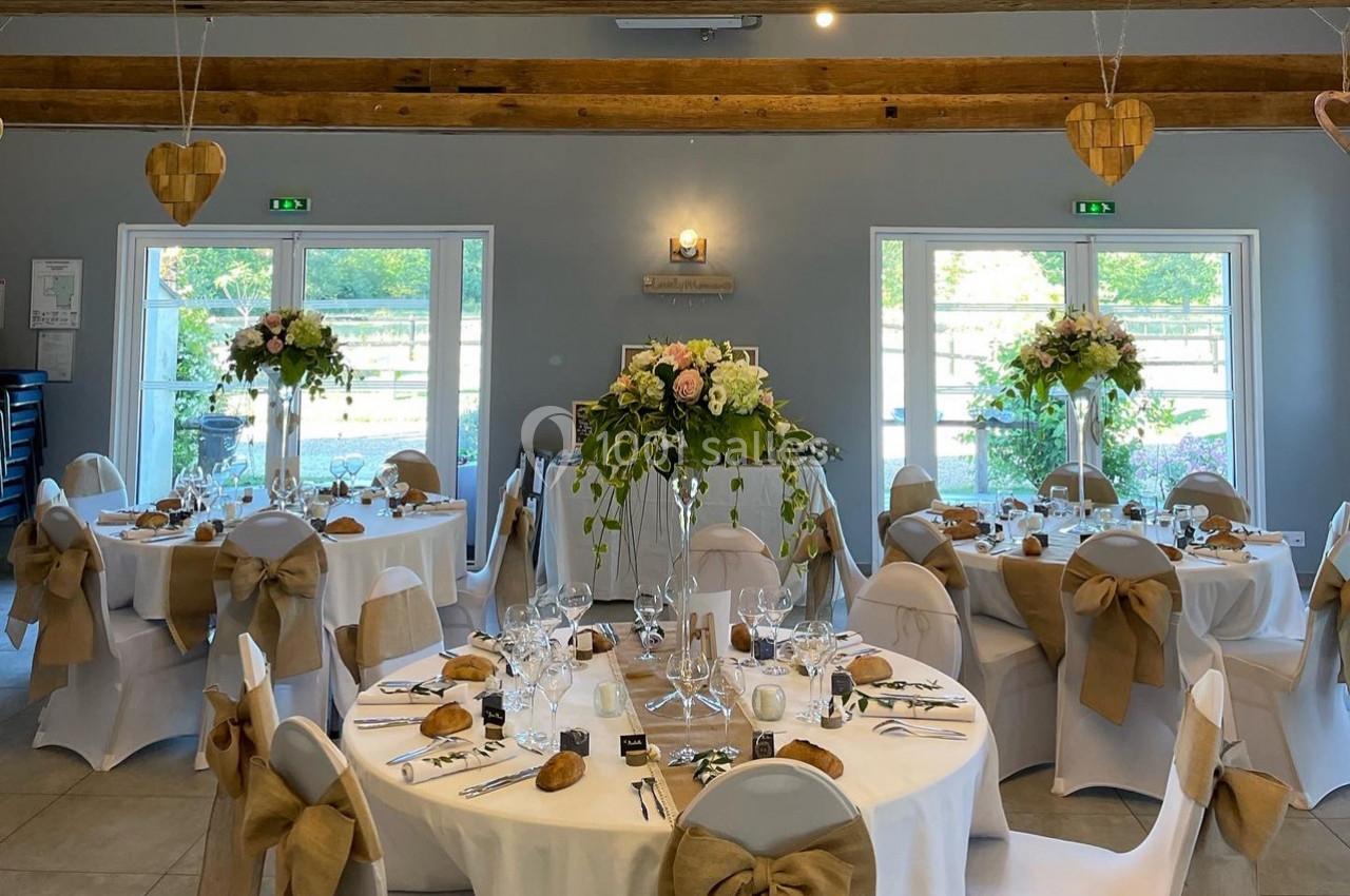 Salle de réception décorée pour un mariage, avec tables rondes dressées, chaises ornées de nœuds en toile de jute et fleurs.