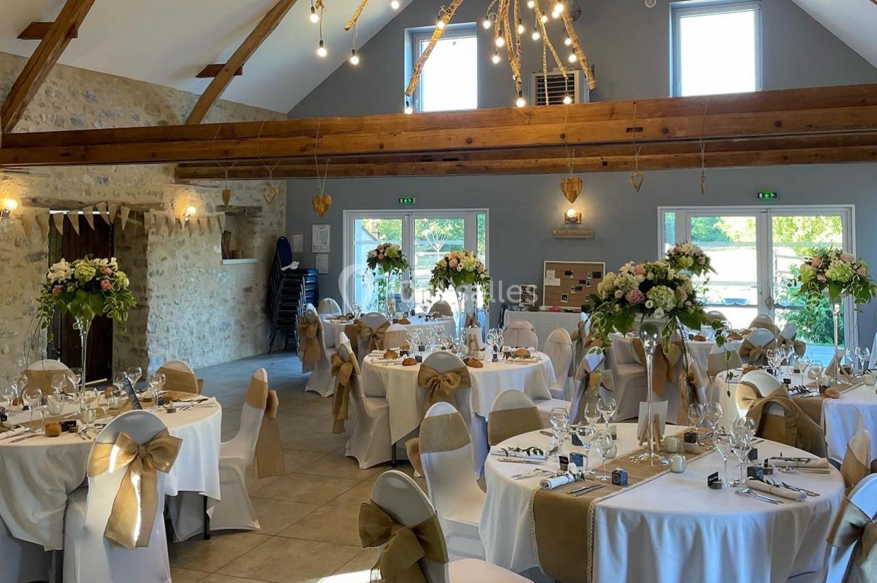Salle de réception décorée pour un mariage, avec tables rondes, nappes blanches, chaises ornées de nœuds et compositions…