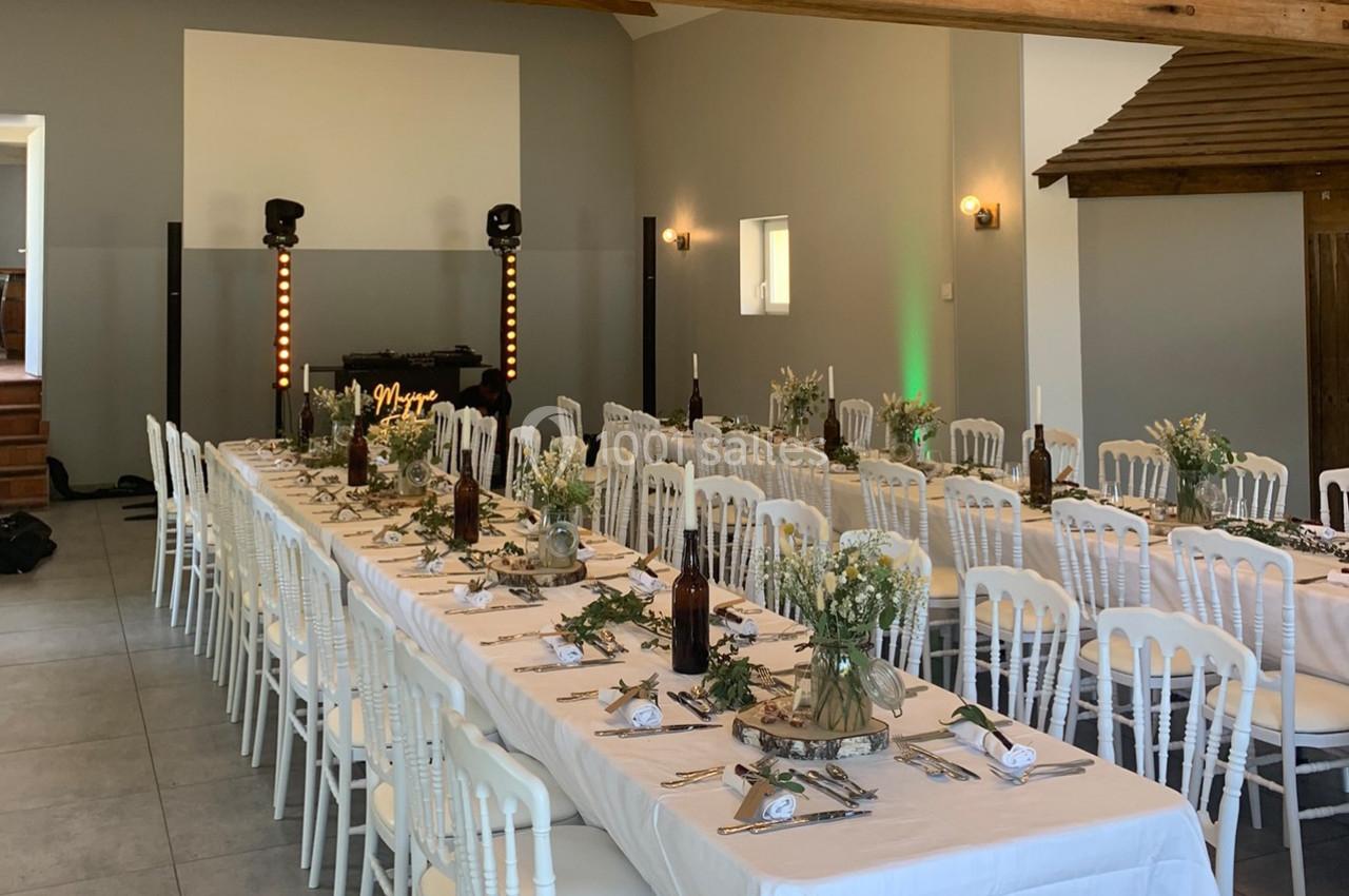 Salle décorée pour un repas festif avec longues tables dressées, chaises blanches et décoration florale.