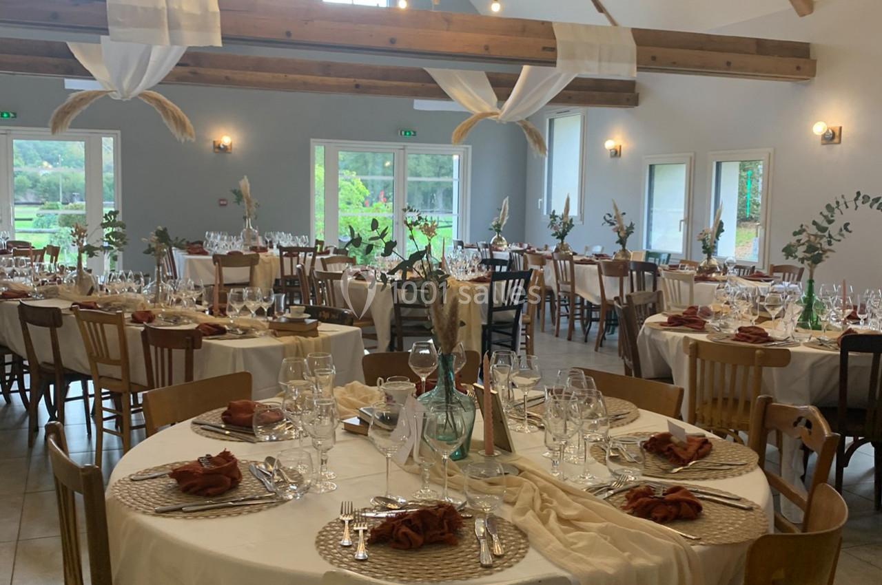 Salle de réception décorée avec des tables rondes dressées, nappes blanches, serviettes marron et éléments végétaux.