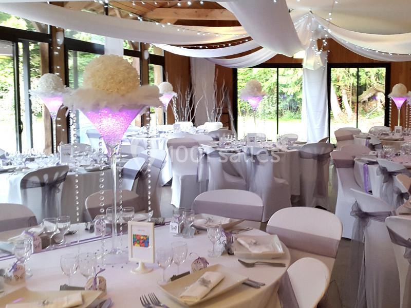 Salle de réception décorée avec des tables dressées, des nappes blanches, des chaises ornées de rubans et des centres de…