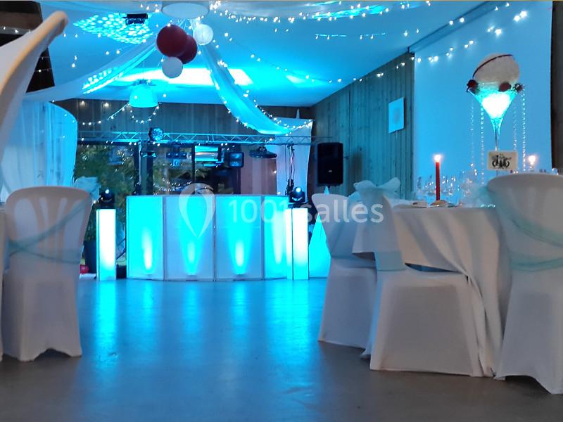 Salle décorée pour un événement avec tables nappées de blanc, guirlandes lumineuses et piste de danse éclairée en bleu.
