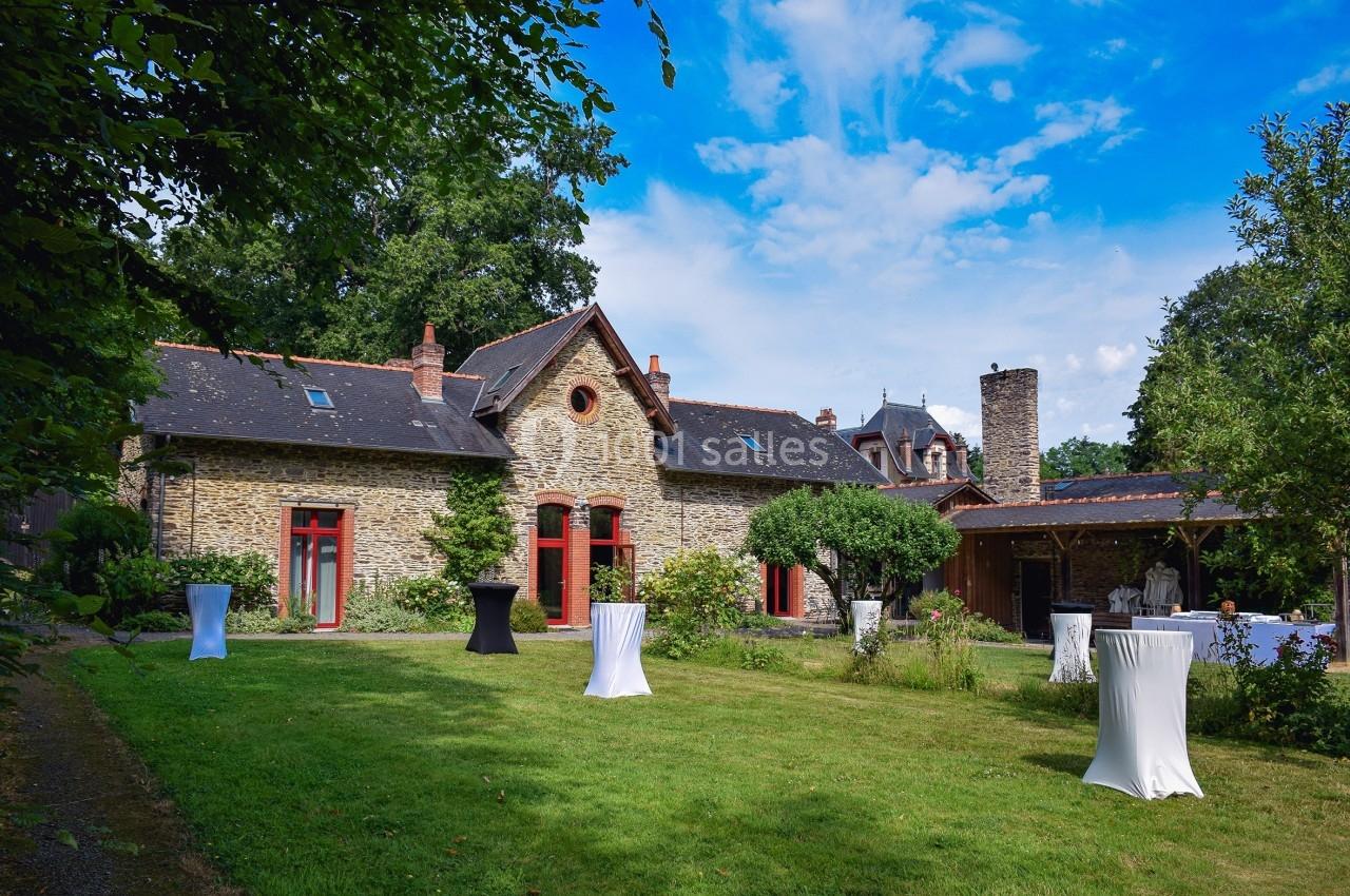 Maison en pierre avec fenêtres rouges entourée d'un jardin verdoyant, tables hautes dressées sur la pelouse.