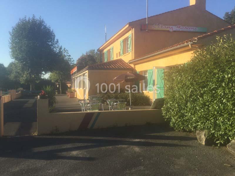 Façade d'un bâtiment jaune avec terrasse extérieure, tables et parasols, entouré de végétation.