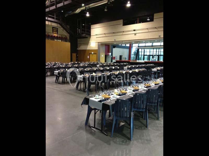 Salle aménagée avec de longues tables dressées pour un repas, chaises alignées et éclairage intérieur.