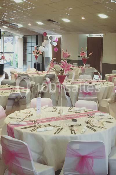 Salle décorée pour un événement avec des tables dressées, nappes blanches, rubans roses et centres de table fleuris.