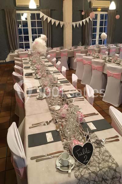 Tables décorées pour un mariage avec nappes blanches, rubans roses, vaisselle et centre de table élégants.