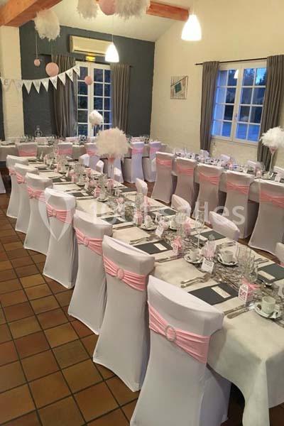 Salle décorée pour un événement avec des tables alignées, nappes blanches, housses de chaise et rubans roses.