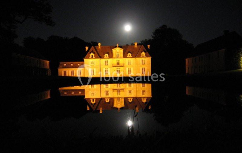 Façade éclairée d'un château se reflétant dans l'eau sous une pleine lune, entouré de bâtiments annexes dans l'obscurité.
